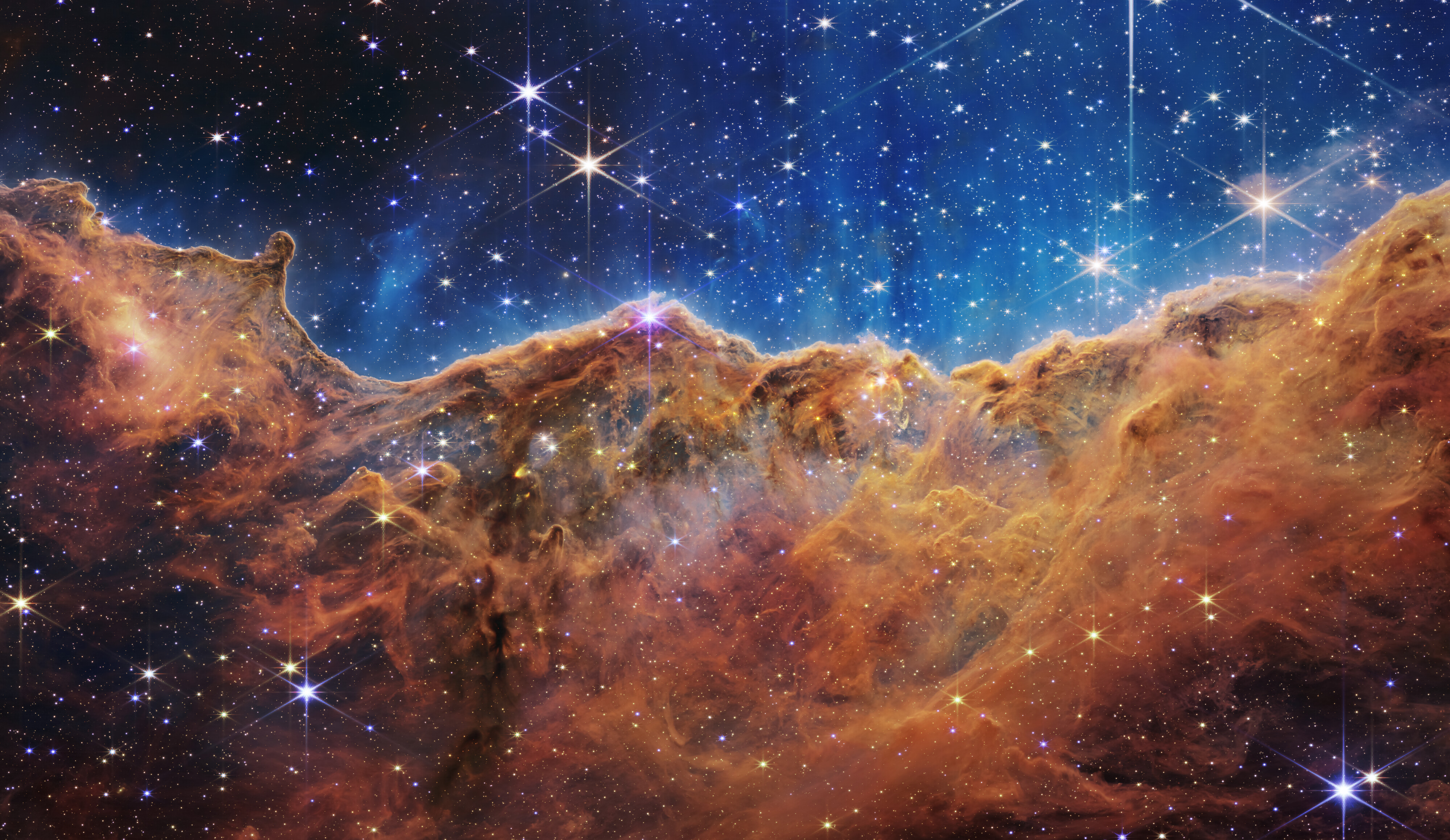Webb telescope view of the Cosmic Cliffs in the Carina Nebula, with amber gas clouds beneath a blue field of stars.